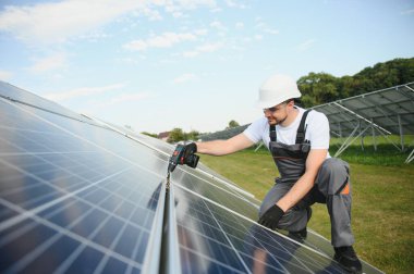 Erkek işçilerin güneş modülleri kurarken ve fotovoltaik güneş dizisini desteklerken yan görünümü. Güneş paneliyle çalışırken kaskını takan bir elektrikçi. Güneş enerjisi kavramı.