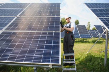 Elinde matkap, elinde tornavida olan üniformalı bir erkek işçi güneş panellerinin yanındaki bir merdiven üzerinde..