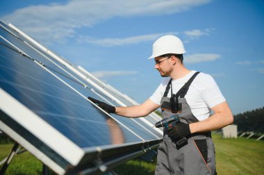 Erkek işçilerin güneş modülleri kurarken ve fotovoltaik güneş dizisini desteklerken yan görünümü. Güneş paneliyle çalışırken kaskını takan bir elektrikçi. Güneş enerjisi kavramı.