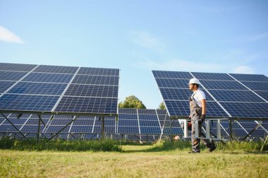 Mavi gökyüzüne karşı güneş panellerinin yanında duran beyaz şapkalı genç Hintli teknisyen portresi. Endüstriyel işçi güneş sistemi kurulumu, yenilenebilir yeşil enerji üretimi kavramı