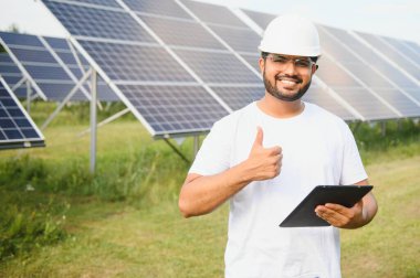 Genç Hintli erkek mühendisin portresi güneş panellerinin yanında duruyor, açık mavi gökyüzü arka planı, yenilenebilir ve temiz enerjisi var. beceri Hindistan, kopyalama alanı.