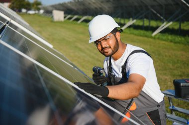Mavi gökyüzüne karşı güneş panellerinin yanında duran beyaz şapkalı genç Hintli teknisyen portresi. Endüstriyel işçi güneş sistemi kurulumu, yenilenebilir yeşil enerji üretimi kavramı