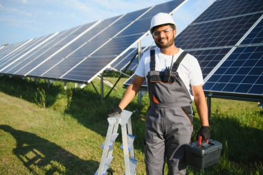 Mavi gökyüzüne karşı güneş panellerinin yanında duran beyaz şapkalı genç Hintli teknisyen portresi. Endüstriyel işçi güneş sistemi kurulumu, yenilenebilir yeşil enerji üretimi kavramı