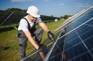 Erkek işçilerin güneş modülleri kurarken ve fotovoltaik güneş dizisini desteklerken yan görünümü. Güneş paneliyle çalışırken kaskını takan bir elektrikçi. Güneş enerjisi kavramı.