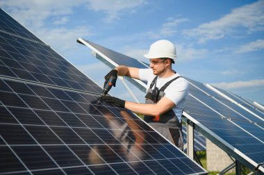 Elinde matkap, elinde tornavida olan üniformalı bir erkek işçi güneş panellerinin yanındaki bir merdiven üzerinde..