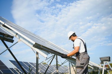 Mavi gökyüzüne karşı güneş panellerinin yanında duran beyaz şapkalı genç Hintli teknisyen portresi. Endüstriyel işçi güneş sistemi kurulumu, yenilenebilir yeşil enerji üretimi kavramı