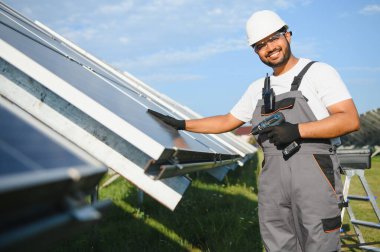 Mavi gökyüzüne karşı güneş panellerinin yanında duran beyaz şapkalı genç Hintli teknisyen portresi. Endüstriyel işçi güneş sistemi kurulumu, yenilenebilir yeşil enerji üretimi kavramı