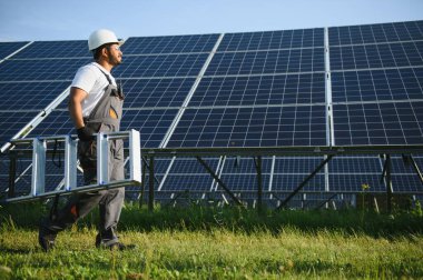 Mavi gökyüzüne karşı güneş panellerinin yanında duran beyaz şapkalı genç Hintli teknisyen portresi. Endüstriyel işçi güneş sistemi kurulumu, yenilenebilir yeşil enerji üretimi kavramı