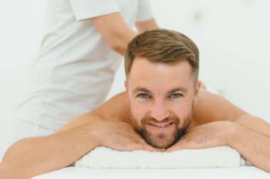 Beyaz üniformalı, uzun saçlı kadın masör gündüz vakti parlak salonda kanepede uzanan adama sırt masajı yapıyor..