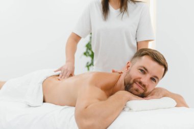 Beyaz üniformalı, uzun saçlı kadın masör gündüz vakti parlak salonda kanepede uzanan adama sırt masajı yapıyor..
