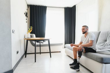 Thoughtful young pensive man amputee with prosthetic leg disability above knee transfemoral leg prosthesis sitting on sofa at home. People with amputation disabilities everyday life. clipart