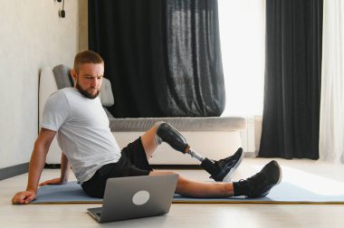 Sporty fit young man amputee with prosthetic leg disability prosthesis doing training exercises on mat at home using laptop. Inclusive online sport classes for people with disabilities concept. clipart