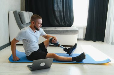 Sportif formda, bacağı sakat, protez protezli bir genç adam evde dizüstü bilgisayar kullanarak antrenman yapıyor. Engelliler için özel online spor dersleri.
