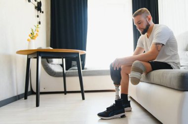 Thoughtful young pensive man amputee with prosthetic leg disability above knee transfemoral leg prosthesis sitting on sofa at home. People with amputation disabilities everyday life. clipart