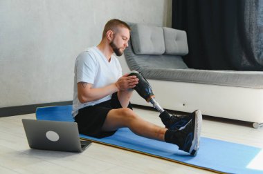Sportif formda, bacağı sakat, protez protezli bir genç adam evde dizüstü bilgisayar kullanarak antrenman yapıyor. Engelliler için özel online spor dersleri.