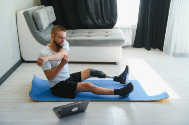 Sportif formda, bacağı sakat, protez protezli bir genç adam evde dizüstü bilgisayar kullanarak antrenman yapıyor. Engelliler için özel online spor dersleri.