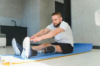 Protez bacaklı bir adamın tam boy portresi evde egzersiz yapıyor ve ayak parmaklarına uzanıyor..