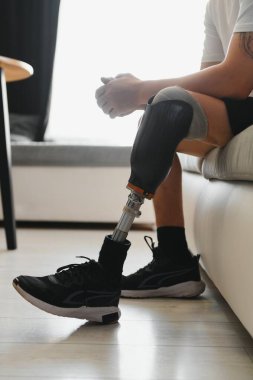 Dizinde protez bacak sakatlığı olan bir erkek bacak protezi takıyor. Kanepede oturuyor. Kapatın. Ampütasyon engelli insanlar günlük yaşam konsepti.