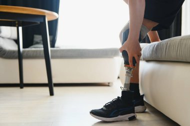 Man amputee with prosthetic leg disability on above knee transfemoral leg prosthesis wearing shoe sitting on sofa, close up. People with amputation disabilities everyday life concept. clipart