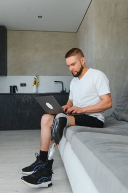 Evde kanepede otururken dizüstü bilgisayar kullanan protez bacaklı yetişkin bir adamın tam boy portresi..