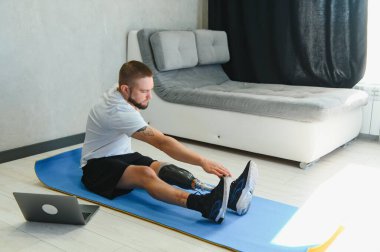 Sportif formda, bacağı sakat, protez protezli bir genç adam evde dizüstü bilgisayar kullanarak antrenman yapıyor. Engelliler için özel online spor dersleri.