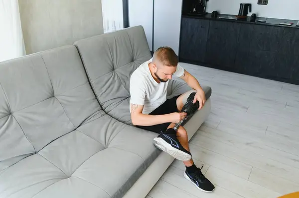 stock image Thoughtful young pensive man amputee with prosthetic leg disability above knee transfemoral leg prosthesis sitting on sofa at home. People with amputation disabilities everyday life.