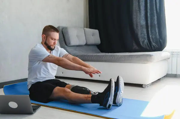 stock image Sporty fit young man amputee with prosthetic leg disability prosthesis doing training exercises on mat at home using laptop. Inclusive online sport classes for people with disabilities concept.