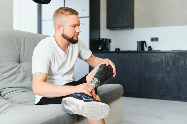 stock image Thoughtful young pensive man amputee with prosthetic leg disability above knee transfemoral leg prosthesis sitting on sofa at home. People with amputation disabilities everyday life.