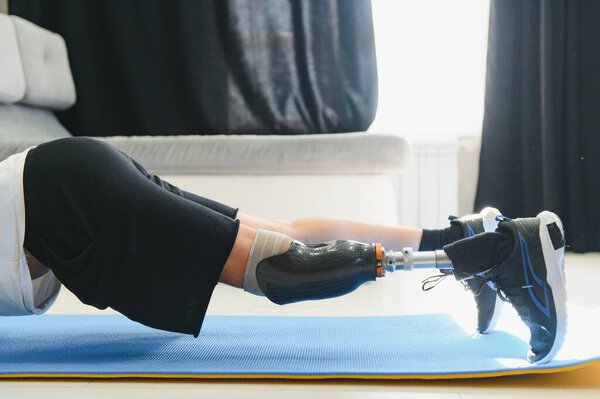 Спортивный молодой человек с ампутированной ногой с протезом протезирования ноги делает упражнения на доске дома на коврике. Концепция эксклюзивной спортивной подготовки для людей с ограниченными возможностями. Подлинный выстрел