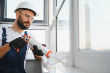 Genç adam, giyinmiş bir üniforma içinde ev onarım yapıyor. Tamirci silikon su kaçağı önlemek için kalafat silahtan uyguluyor.