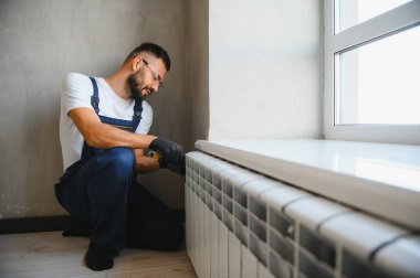 Mavi tulumlu bir tesisatçı radyatör tamir ediyor. Ayarlanabilir bir anahtarla musluğu sıkıyor. Elinde bir alet olan bir çanta..