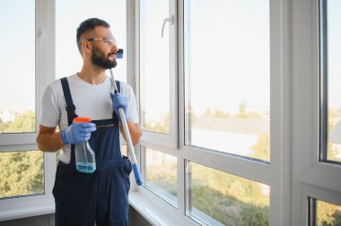Beyaz tenli yakışıklı bir adam, temizlikçi, mavi tulumlu, kepli, sidikli, pencereleri temizleyen, özel paspas kullanan, işinden zevk alan. Yüksek kalite fotoğraf.