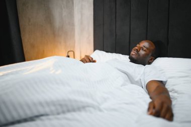 Afro-Amerikalı bir adam rahat beyaz bir yatakta uyuyor güzel güzel güzel rüyalar görüyor, sakin melez bir erkek rahat bir yatak odasında çarşaf örtüsünün altında uyukluyor..