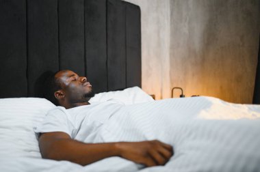 Afro-Amerikalı bir adam rahat beyaz bir yatakta uyuyor güzel güzel güzel rüyalar görüyor, sakin melez bir erkek rahat bir yatak odasında çarşaf örtüsünün altında uyukluyor..