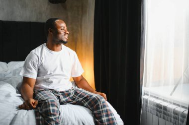 Tamamen dinlenmiş Afro-Amerikalı bir adamın sabah uyandıktan sonra pijama giyip yatağa oturması, pencereye bakması, vücudunu esnetmesi, güneş ışığı, fotokopi alanı. Rahat, sağlıklı uyku konsepti
