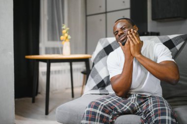 Afro-Amerikalı bir adam evinde, koltukta şiddetli diş ağrısı çekiyor. Elini yanağına dayayıp acı içinde yüzünü buruşturuyor..