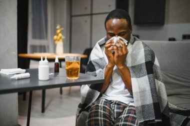 Evdeki kanepede sıcak bir battaniyenin altında yatıp kağıt mendile hapşıran hasta, Afro-Amerikan bir adam. Soğuk algınlığı, grip, mevsimsel kış enfeksiyonları