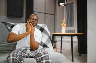 Afro-Amerikalı bir adam evinde, koltukta şiddetli diş ağrısı çekiyor. Elini yanağına dayayıp acı içinde yüzünü buruşturuyor..