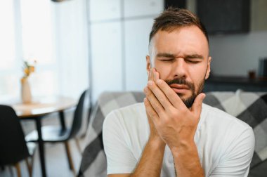 Diş ağrısı. Mutsuz, üzgün, üzgün, beyaz bir adam evdeki kanepede oturuyor, elini yanağına yaklaştırıyor, yüzünü buruşturuyor, dişleri ağrıyor, bir dişçiye görünmesi gerekiyor..