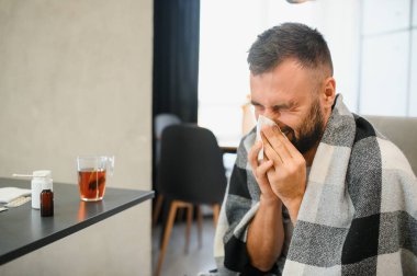 Sakallı, hasta, üzgün bir adam evdeki kanepede oturuyor. Burun nezlesi salgını, koronavirüs salgını, çoban salgını, hapşırma....