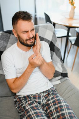 Diş sağlığı sorunları konsepti. Güçlü diş ağrısı çeken bir adam. İltihaplı sinirleri, diş apsesi ya da diş hassasiyeti olan üzgün bir erkek hasta kanepeye oturmuş ve acı içinde yanaklarına dokunuyor..