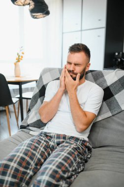 Diş sağlığı sorunları konsepti. Güçlü diş ağrısı çeken bir adam. İltihaplı sinirleri, diş apsesi ya da diş hassasiyeti olan üzgün bir erkek hasta kanepeye oturmuş ve acı içinde yanaklarına dokunuyor..