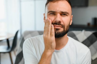 Diş sağlığı sorunları konsepti. Güçlü diş ağrısı çeken bir adam. İltihaplı sinirleri, diş apsesi ya da diş hassasiyeti olan üzgün bir erkek hasta kanepeye oturmuş ve acı içinde yanaklarına dokunuyor..