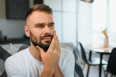 Diş sağlığı sorunları konsepti. Güçlü diş ağrısı çeken bir adam. İltihaplı sinirleri, diş apsesi ya da diş hassasiyeti olan üzgün bir erkek hasta kanepeye oturmuş ve acı içinde yanaklarına dokunuyor..