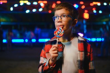 Akşamları eğlence parkında ya da sirkte renkli bir lolipop yiyen beyaz bir çocuk..