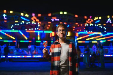 Akşamları eğlence parkında ya da sirkte renkli bir lolipop yiyen beyaz bir çocuk..