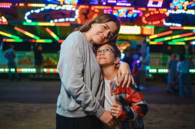 Sarışın, kıvırcık saçlı ve annesi renkli bir lolipopu yiyen sevimli bir çocuk bir atlıkarıncanın arka planında duruyor.