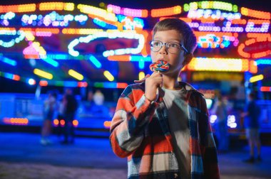 Akşamları eğlence parkında ya da sirkte renkli bir lolipop yiyen beyaz bir çocuk..