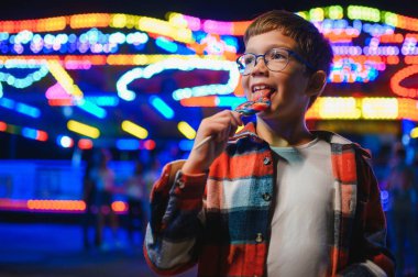 Akşamları eğlence parkında ya da sirkte renkli bir lolipop yiyen beyaz bir çocuk..