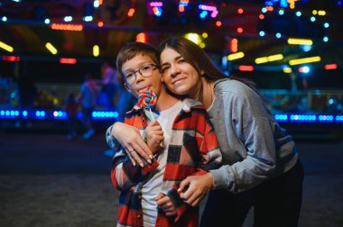 Sevimli bir çocuk ve annesi renkli bir lolipop yiyorlar. Atlıkarıncanın arka planında duruyorlar.